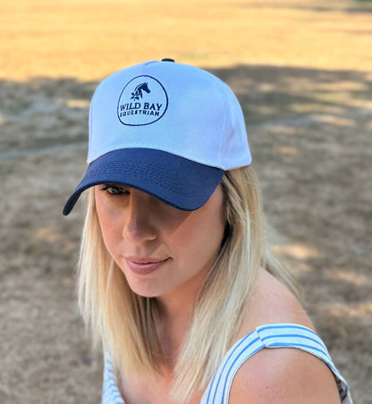 Woman wearing a white and navy Wild Bay Equestrian baseball cap with embroidered logo, outdoors in a sunny field.
